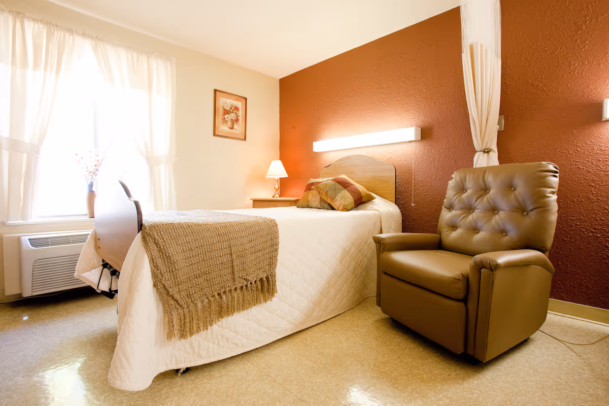 A cozy bedroom in a senior living facility with a single bed covered in a white quilt and a beige throw blanket. There are two decorative pillows on the bed. Next to the bed is a wooden nightstand with a lamp. A large brown leather armchair is positioned beside the bed. The walls are painted beige and dark red, and a framed floral picture hangs on the beige wall. A window with sheer white curtains lets in natural light.