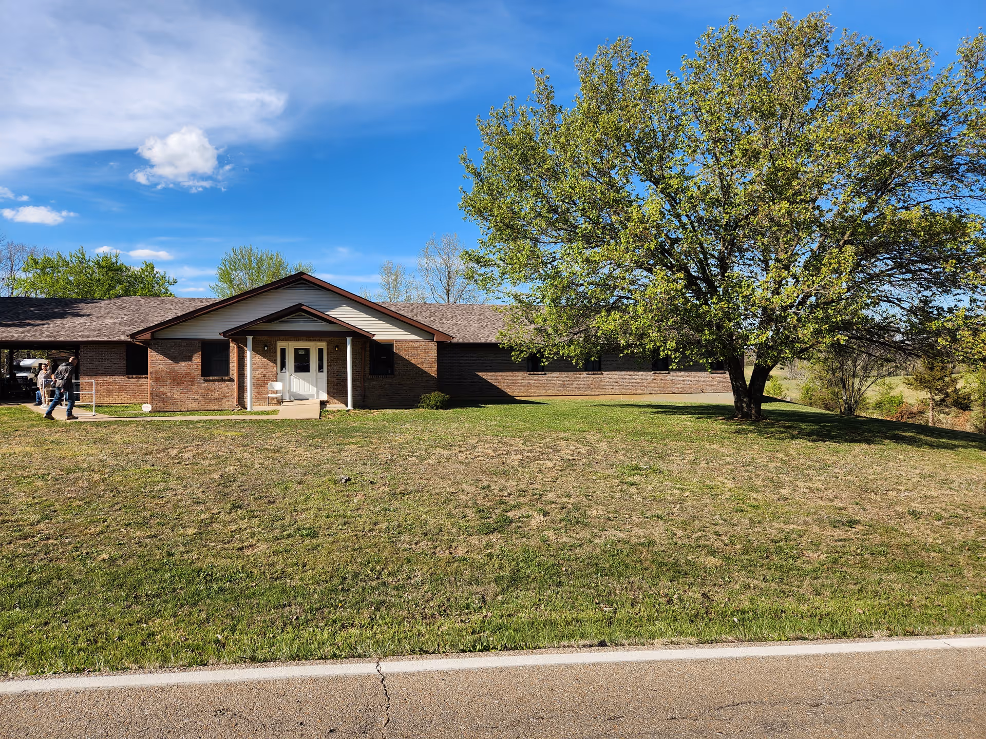 Single-story brick building with a central entrance, large grassy front yard and a big tree under a blue sky.