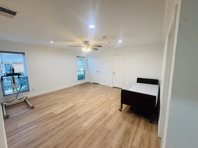 A sparsely furnished bedroom at Evergreen Assisted Living with a bed, exercise equipment, and windows.
