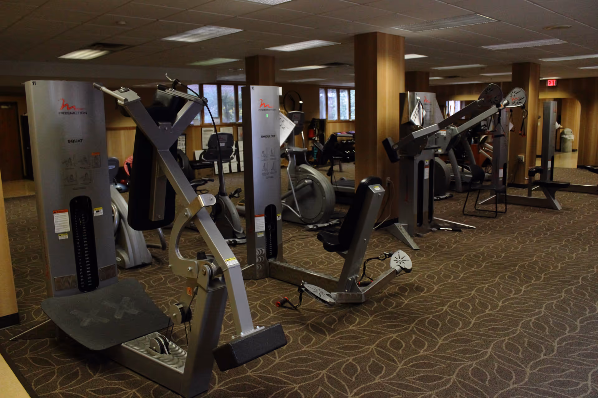 Interior view of a fitness room with multiple exercise machines including squat and shoulder workout equipment, carpeted floor with a leaf pattern, wood-paneled walls, and fluorescent ceiling lights.