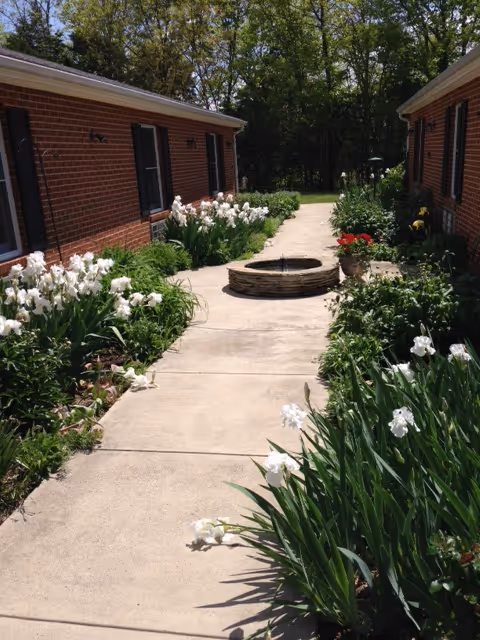 A concrete walkway between two single-story brick buildings with black shutters, bordered by lush green plants and white flowers. At the center of the walkway is a circular stone water fountain. Trees and greenery are visible in the background under a clear sky.