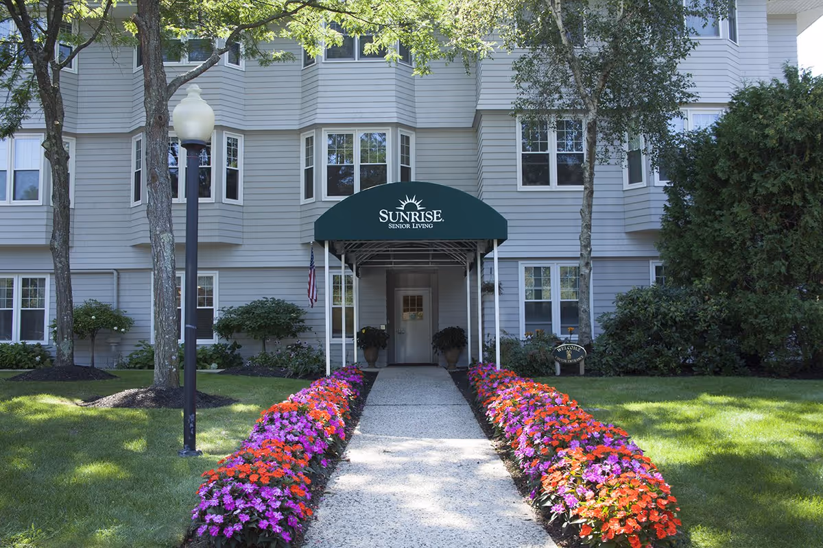 Front entrance of a Sunrise Senior Living building with a green awning and a flower-lined walkway.