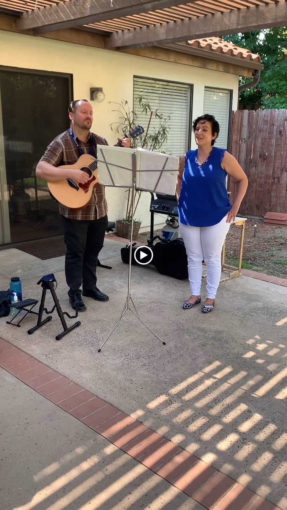 A man playing an acoustic guitar and a woman standing beside him singing outdoors on a patio area with a music stand in front of them. They are under a pergola attached to a building with windows and a wooden fence in the background.
