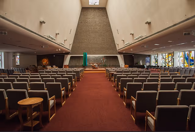 Interior view of a large chapel or auditorium with rows of cushioned chairs arranged on either side of a central aisle covered with a red carpet. The front features a raised platform with religious symbols and decorations, including a cross and a lectern. The walls are high and angled, with stained glass windows visible on the sides.
