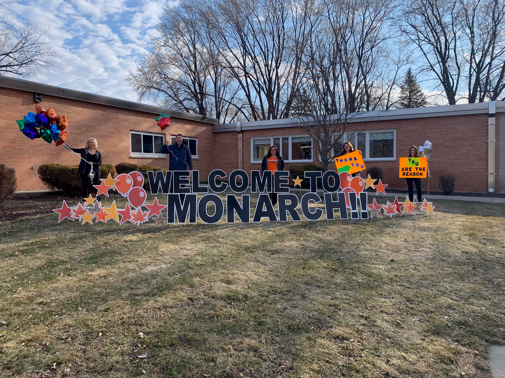 Group of adults standing with a large Welcome to Monarch sign outside a brick building.