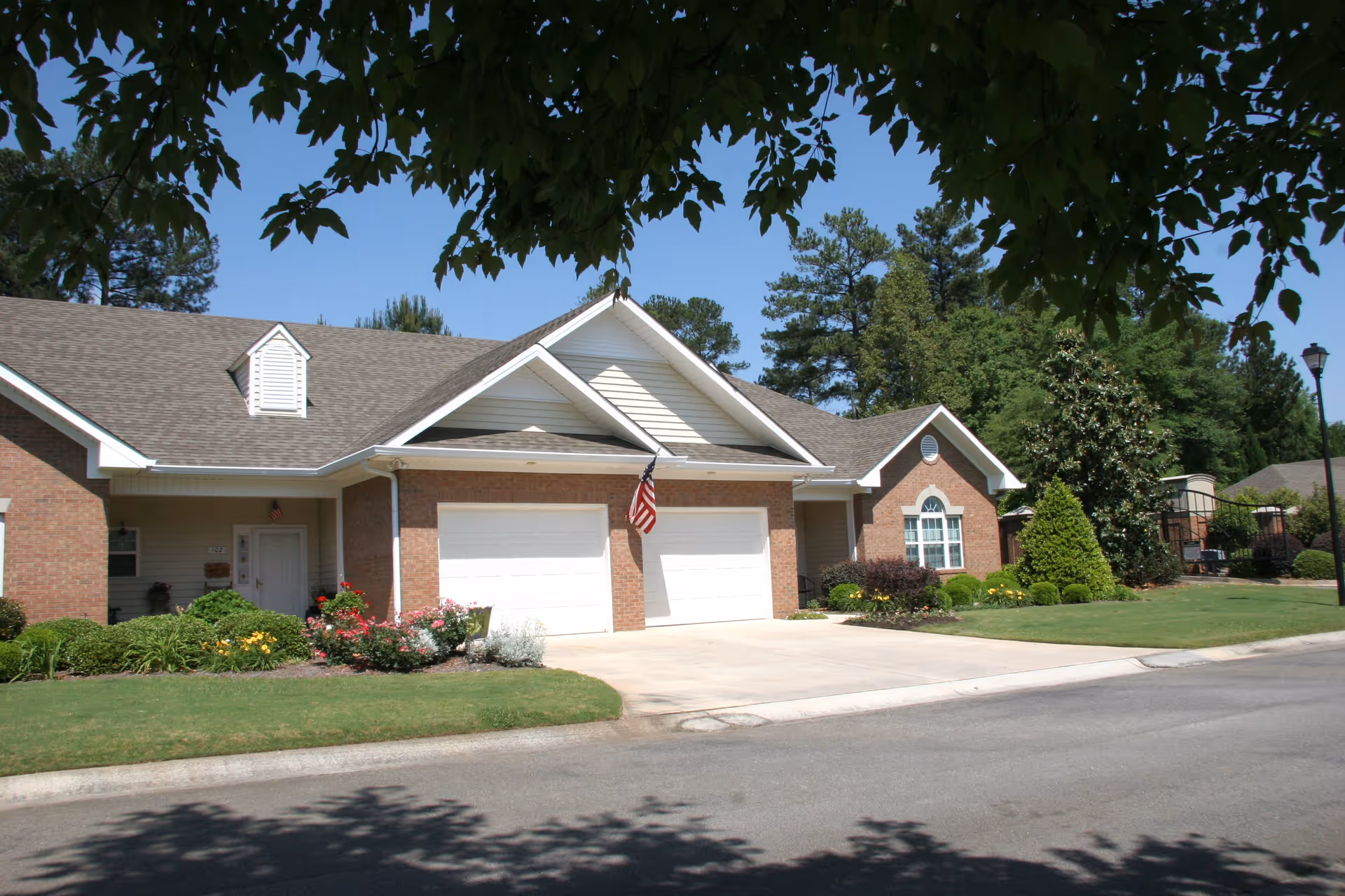 Exterior view of a single-story residential building with a double garage, an American flag mounted between the garage doors, well-maintained lawn, shrubs, and trees under a clear blue sky.