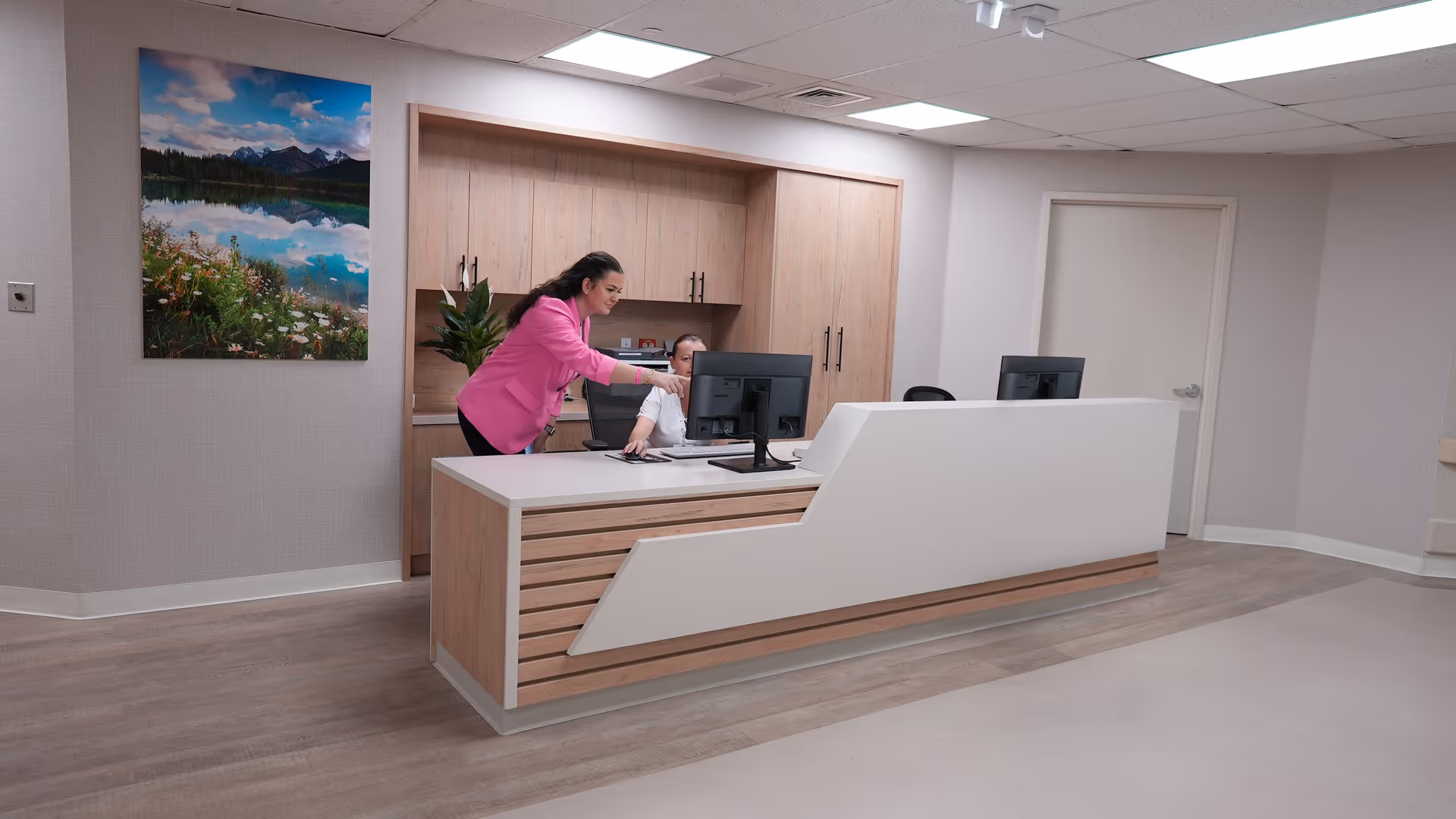 A modern reception area in a nursing and rehabilitation center with a wooden and white reception desk. Two women are interacting at the desk, one standing and pointing at a computer screen while the other is seated. The room has light-colored walls, wood flooring, a large landscape photograph on the wall, and built-in wooden cabinets behind the desk.