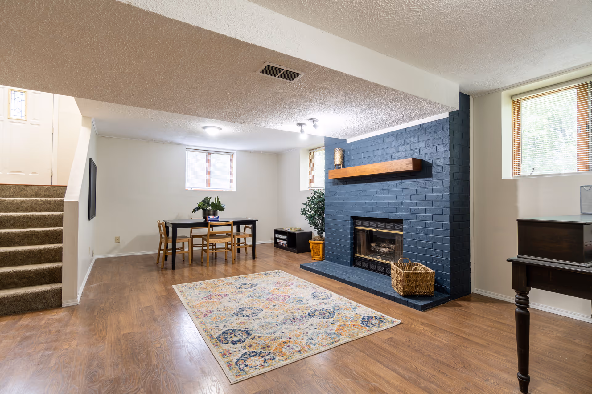 Cozy basement-style living room with blue brick fireplace, dining table, rug, and piano.