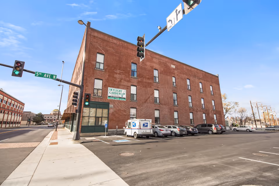 Three-story brick building labeled 'Skylight Gardens' at a street corner with traffic lights and parked cars.