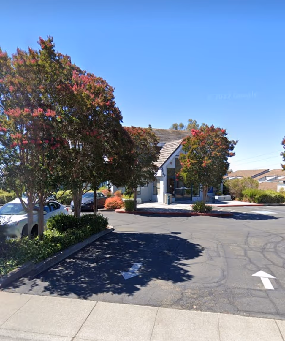 Exterior view of a small building’s front with a parking lot and trees.