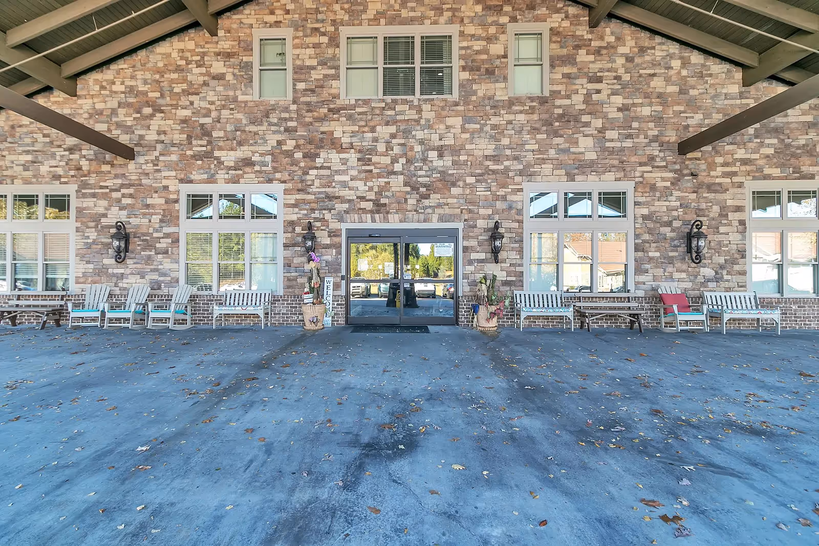 Covered entrance area of a building with stone brick exterior walls, multiple windows, and a glass double door in the center. There are several white benches and chairs arranged along the wall, with some potted plants and decorative lantern-style wall lights. The ground is paved and scattered with fallen leaves.