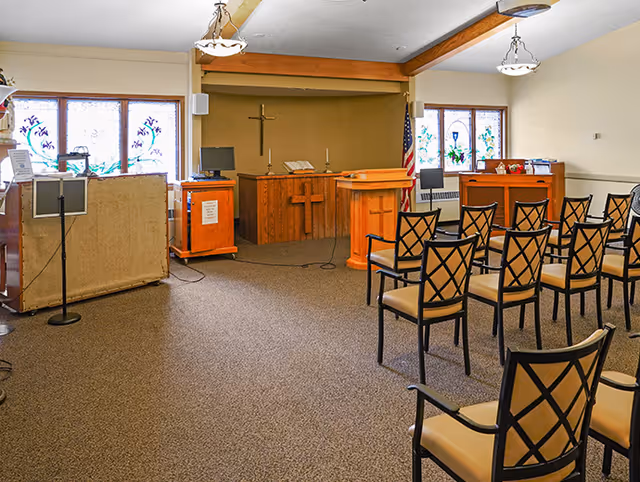 Interior chapel in a senior facility with a wooden pulpit, cross, pews, and flag.