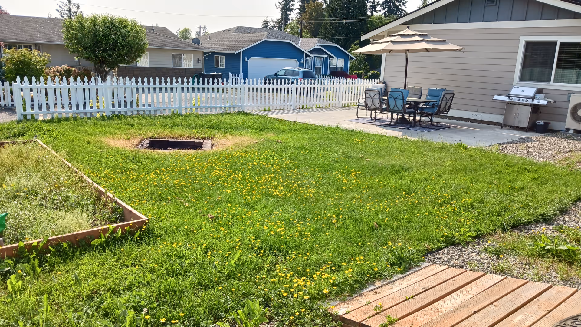 A grassy backyard with a white picket fence, patio dining set, grill, and raised garden bed.