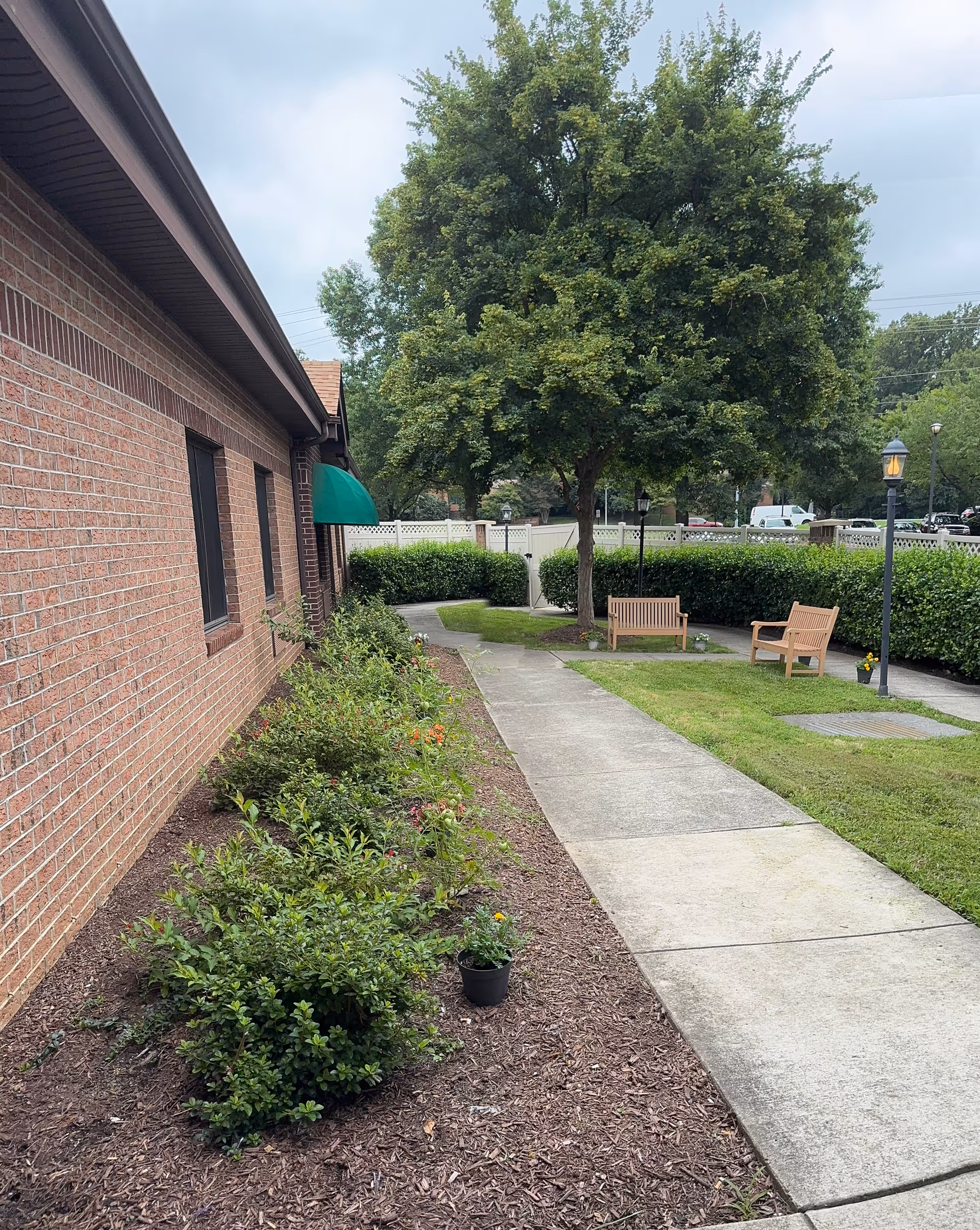 Brick exterior building with a walkway, garden bed, tree, and benches in an outdoor area.