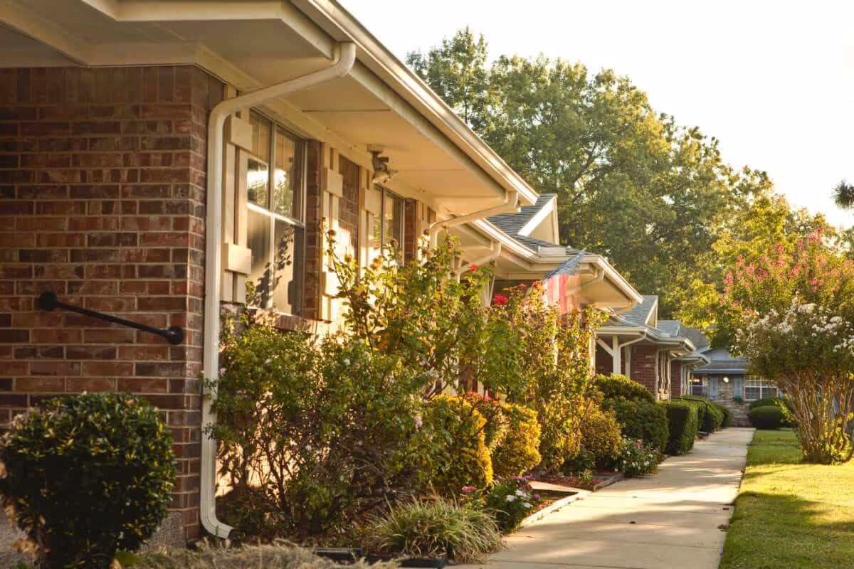 Row of brick senior living buildings with landscaped shrubs along a sunlit walkway.