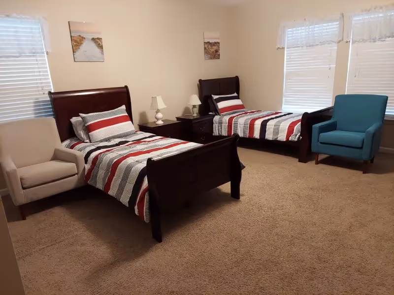 A bedroom with two single beds featuring dark wooden frames and striped bedding in red, white, and black. Each bed has a pillow and is separated by a nightstand with two white lamps. There is a beige armchair next to the left bed and a blue armchair next to the right bed. The room has beige carpet, light-colored walls, two windows with blinds and sheer curtains, and two framed pictures on the walls.