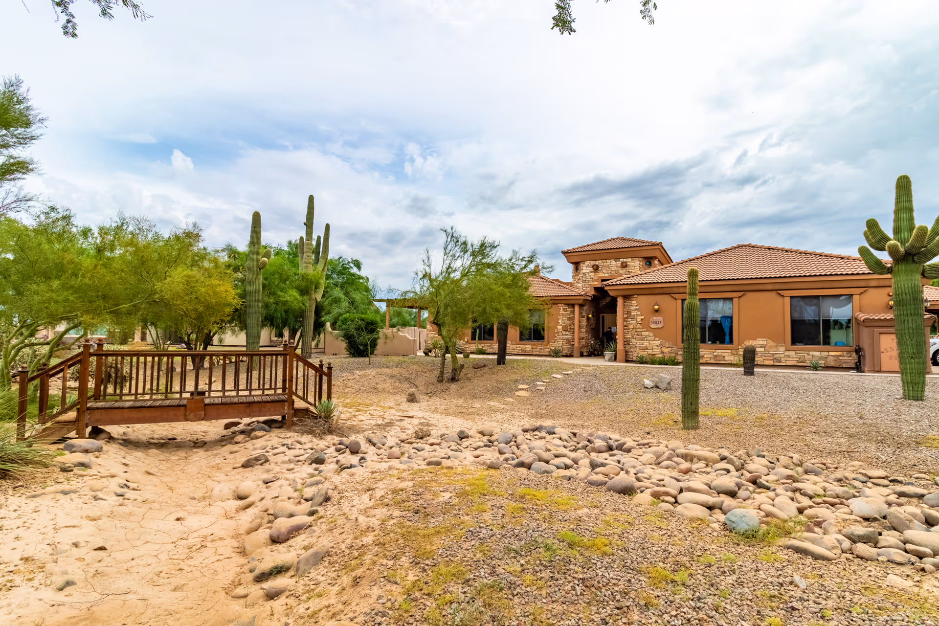 Desert landscaping with cacti and a wooden bridge leads to a tan, stone-accented assisted living building.