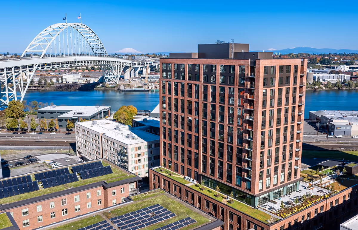 Aerial view of a multi-story brick residential building by a river with a large arched bridge in the background.