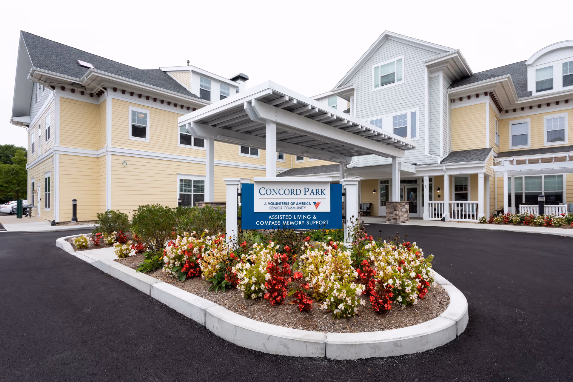 Entrance of Concord Park Senior Living showing a covered porte-cochere, a sign, and a landscaped flower bed in front of a yellow multi-story building.