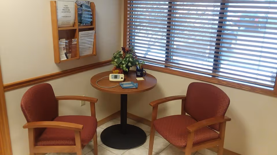 Two red upholstered chairs surround a round table by a window with blinds and a plant.