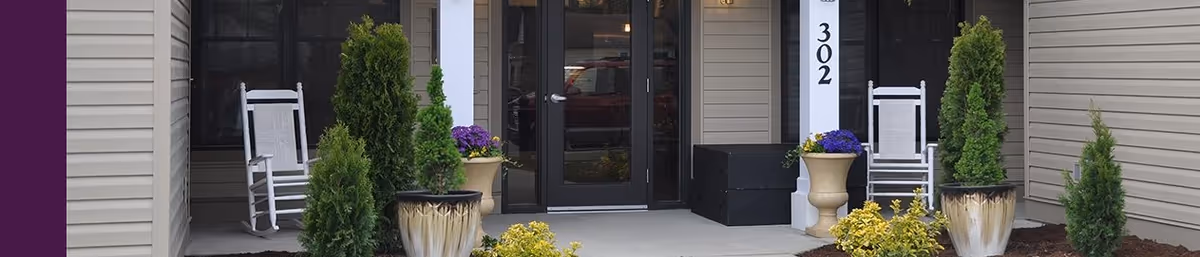 Front entrance of a building with double glass doors, two white rocking chairs on either side of the entrance, potted plants including small evergreen trees and flowers, and the number 302 displayed on a white column.