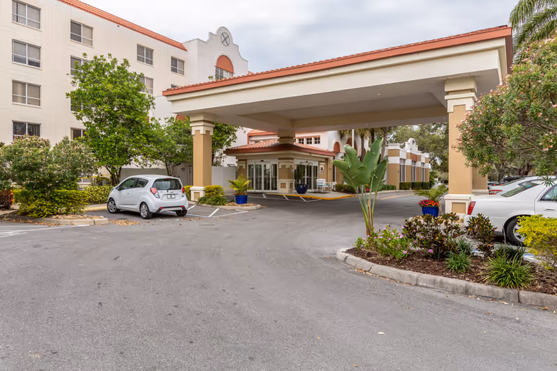 Covered porte-cochere entrance of a senior living building with parked cars and landscaped grounds.
