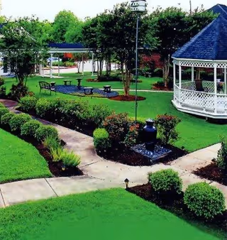 A well-maintained outdoor garden area with green grass, trimmed bushes, a paved walkway, benches, a gazebo with white latticework, and several trees providing shade.