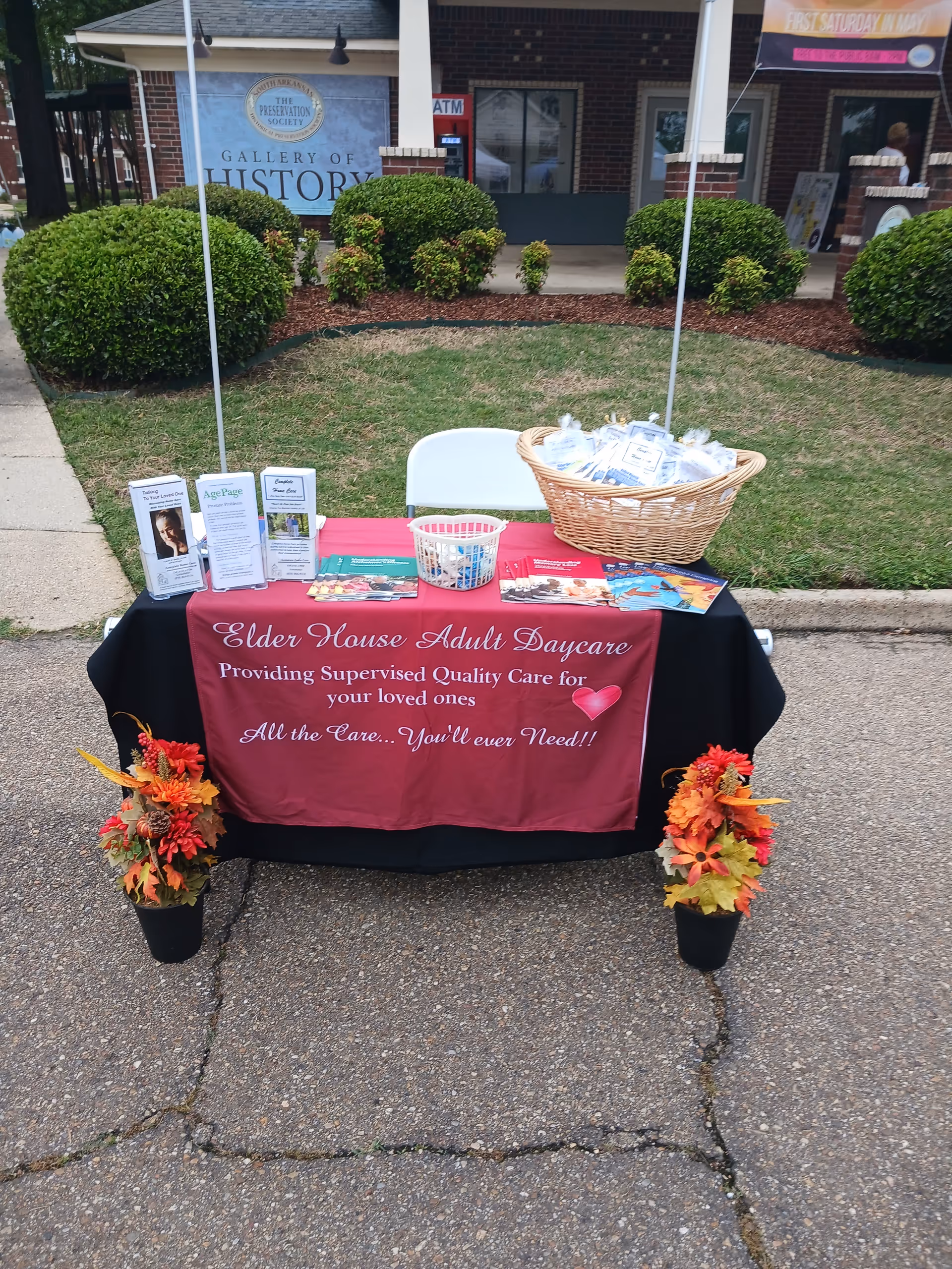 Outdoor promotional table with a maroon banner, brochures, a basket, and fall flowers.