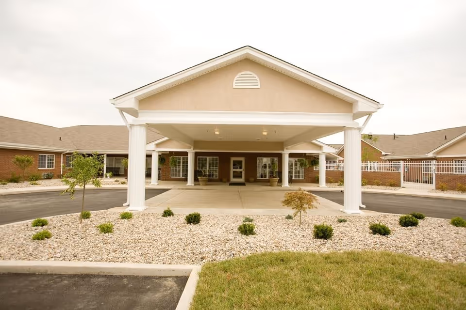 Front exterior view of Stonegate Health Campus featuring a covered entrance with white columns, a driveway, small landscaped areas with bushes and trees, and a beige building with multiple windows and a sloped roof.