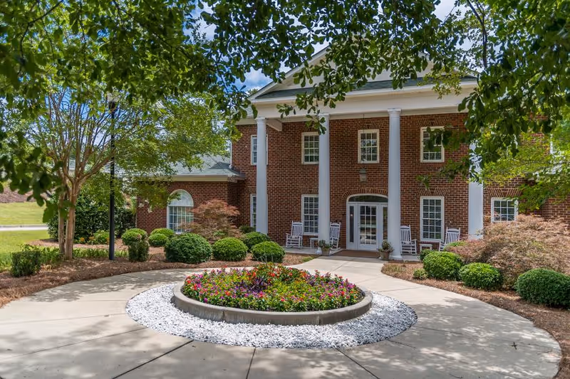 Brick two-story building with white columns, a circular flowerbed in the driveway, and rocking chairs at the entrance.