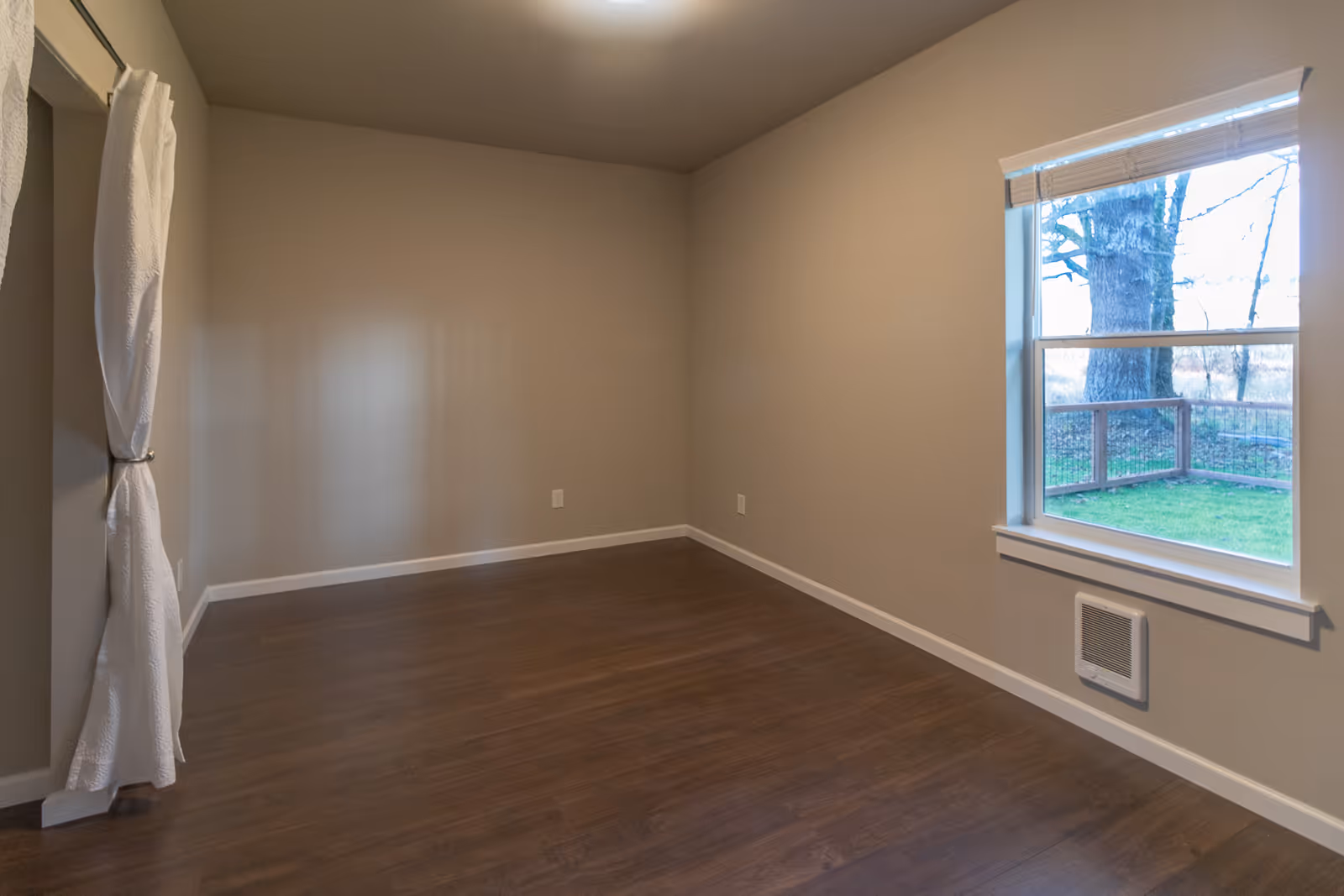 Empty beige interior room with dark wood flooring, window, and curtain on the left.