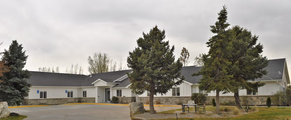 Exterior view of Oak Ridge Assisted Living of Bountiful, showing a single-story building with white siding and stone accents, a dark roof, and a driveway in front. There are several large evergreen trees and a small seating area with a table and chairs on the lawn.