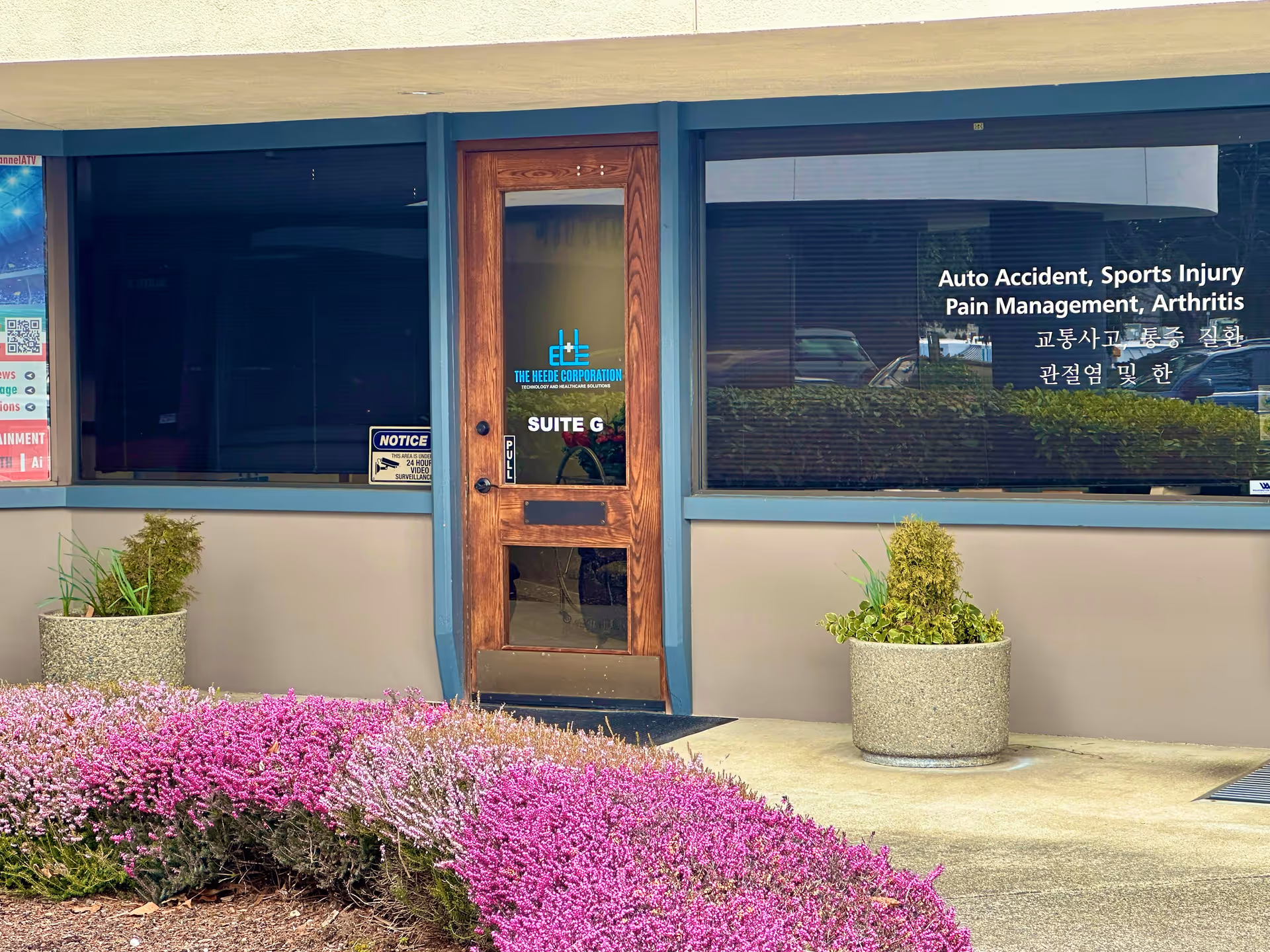 Exterior building entrance with wooden door, large windows, and flowering planters in front.
