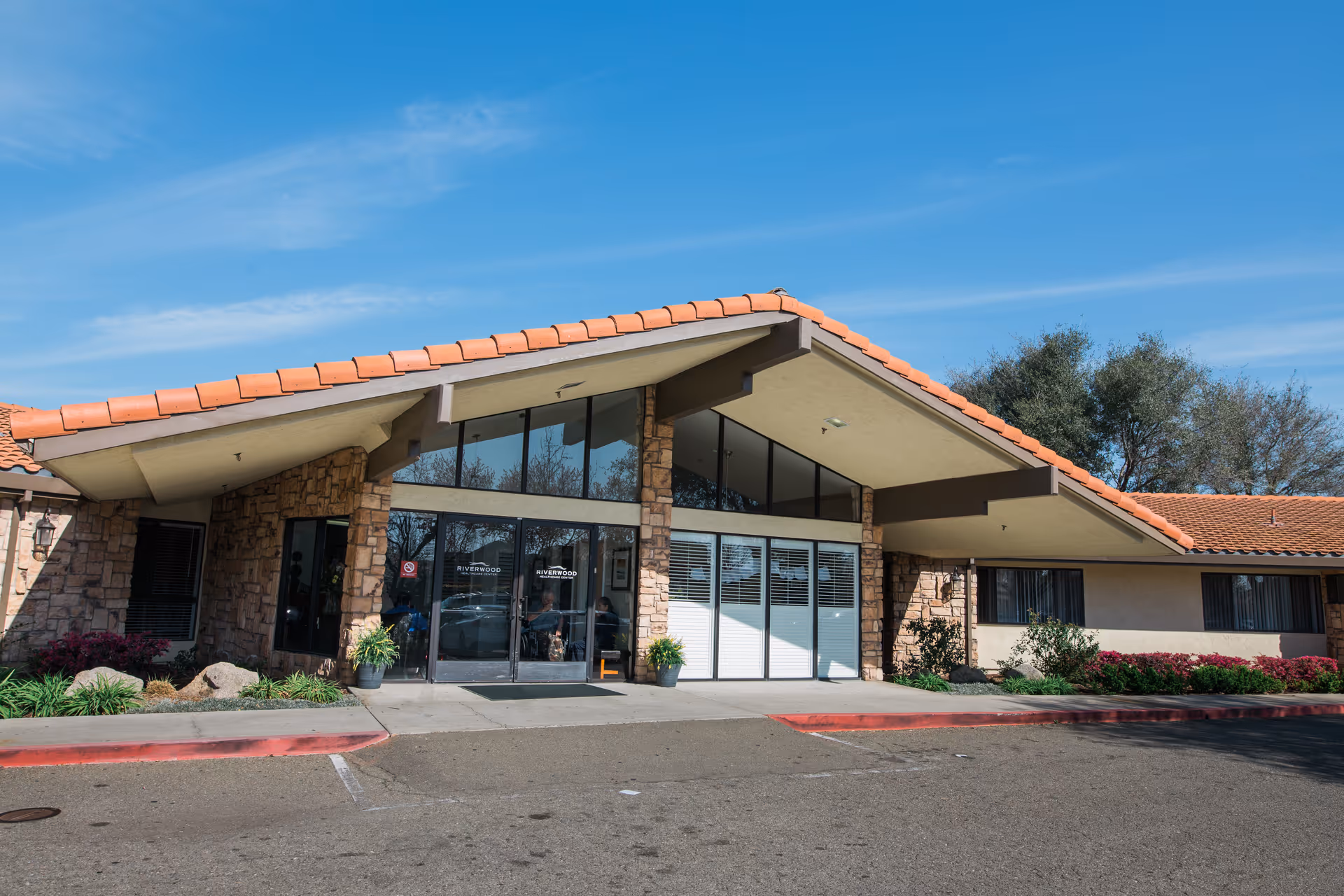 Exterior view of Riverwood Healthcare Center featuring a building with a sloped roof with orange tiles, stone and stucco walls, large glass entrance doors, and surrounding landscaping with bushes and plants under a clear blue sky.