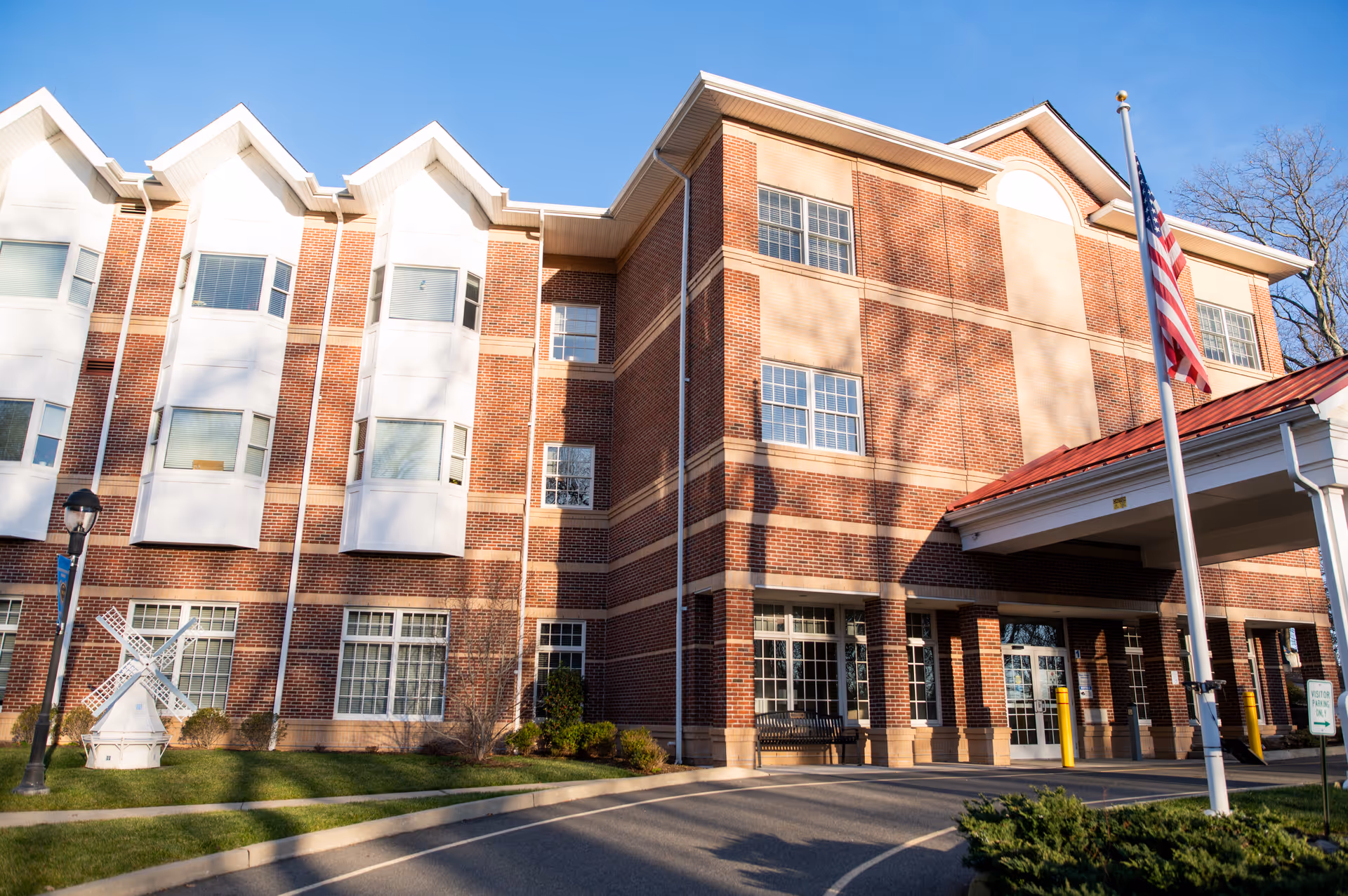 Front entrance of a three-story brick senior living building with a covered drive, American flag, and a small decorative windmill on the lawn.