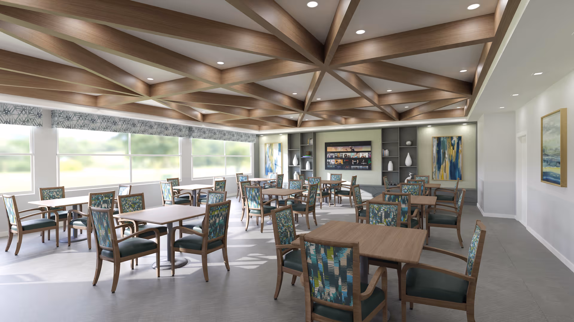A bright and spacious dining room with multiple wooden tables and chairs featuring patterned upholstery. The room has large windows allowing natural light to fill the space, a decorative wooden ceiling with recessed lighting, and modern artwork on the walls. Shelves with decorative items are visible at the far end of the room.