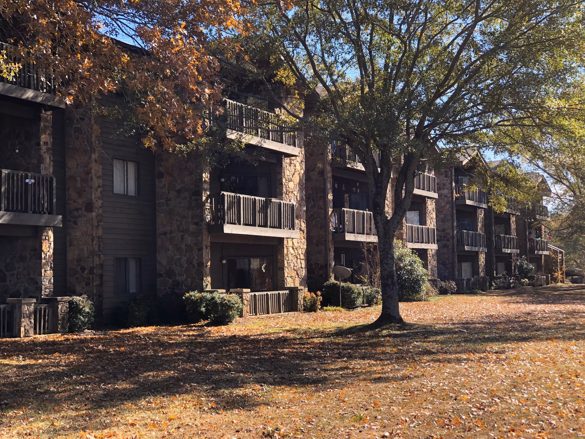Exterior view of a multi-story residential building with stone and wood facade, balconies, and surrounded by trees with autumn leaves on the ground at Indian Rock Village.