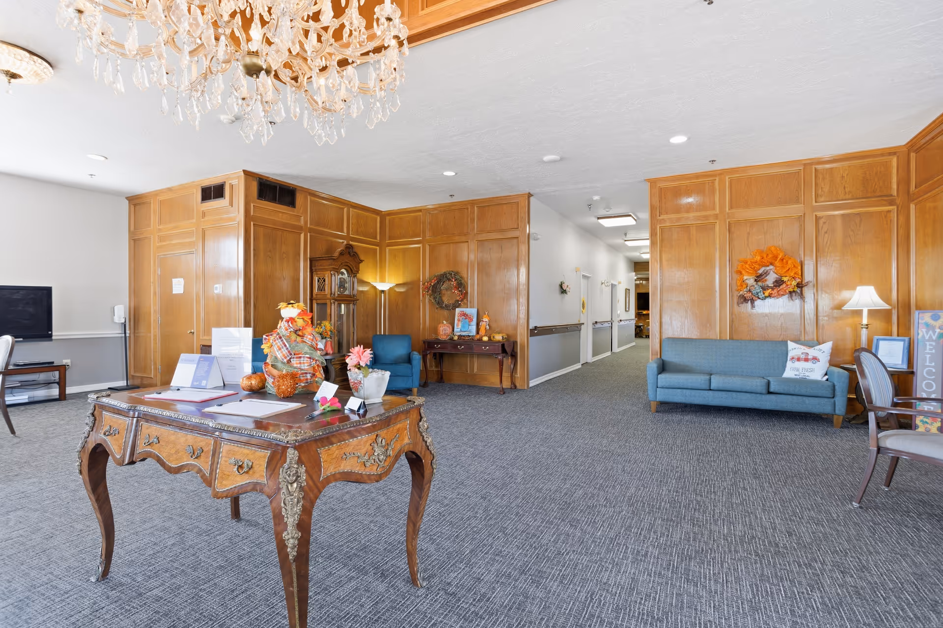 Luxurious senior living lobby with ornate desk, blue sofas, wood paneling, and chandelier.