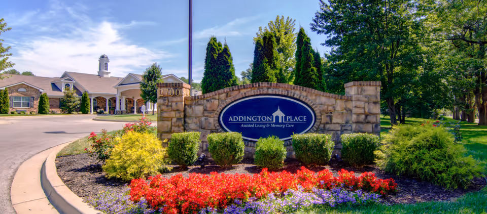 Entrance sign for Addington Place Assisted Living & Memory Care surrounded by colorful flowers and greenery, with the facility building visible in the background under a blue sky.