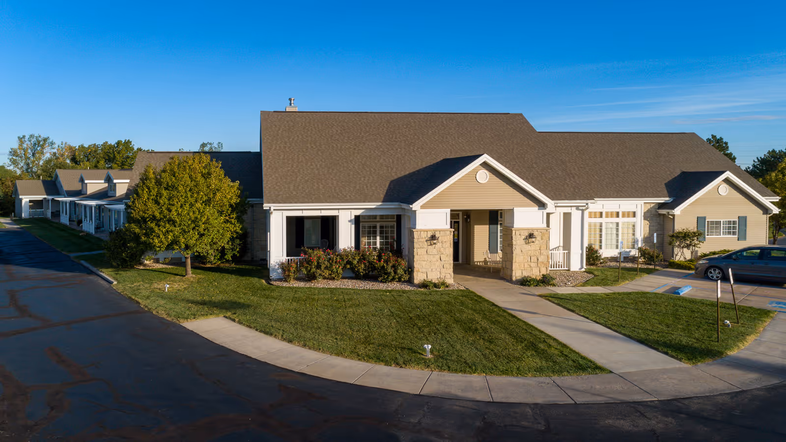 Exterior view of Homestead Assisted Living of Hays, showing a single-story building with a pitched roof, beige siding, stone accents, a covered entrance, and surrounding green lawn with a tree and bushes under a clear blue sky.