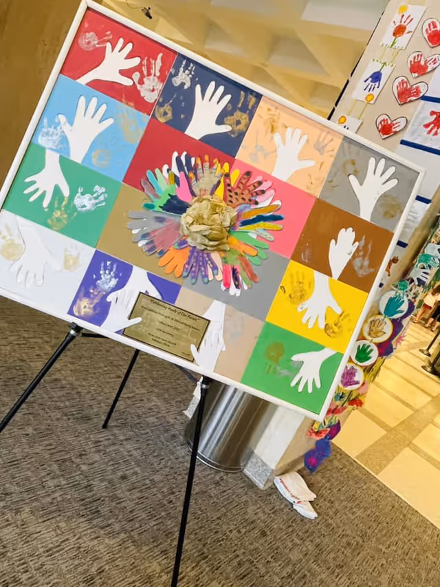 Colorful handprint art board displayed on an easel in a facility lobby interior.