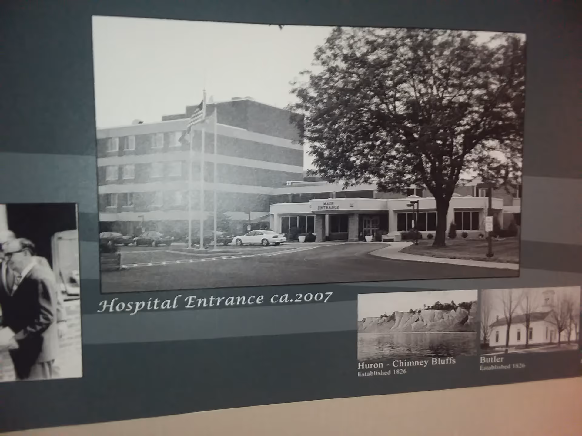 Black-and-white image of a hospital exterior with main entrance, flagpoles, and surrounding parking.