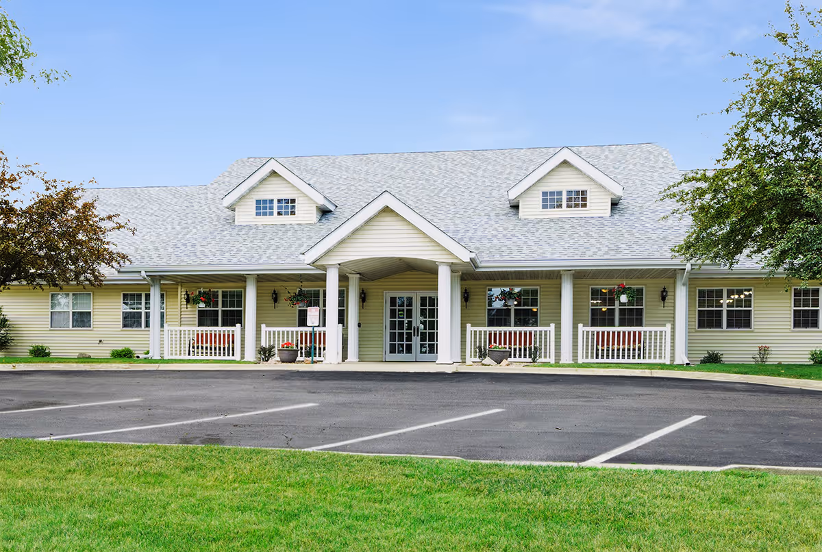Front exterior view of a single-story senior living facility building with a light gray roof, cream-colored siding, white columns, and a covered entrance. There are windows with flower pots and a small porch area on either side of the entrance. The building is surrounded by green grass and trees, with a paved parking area in front.