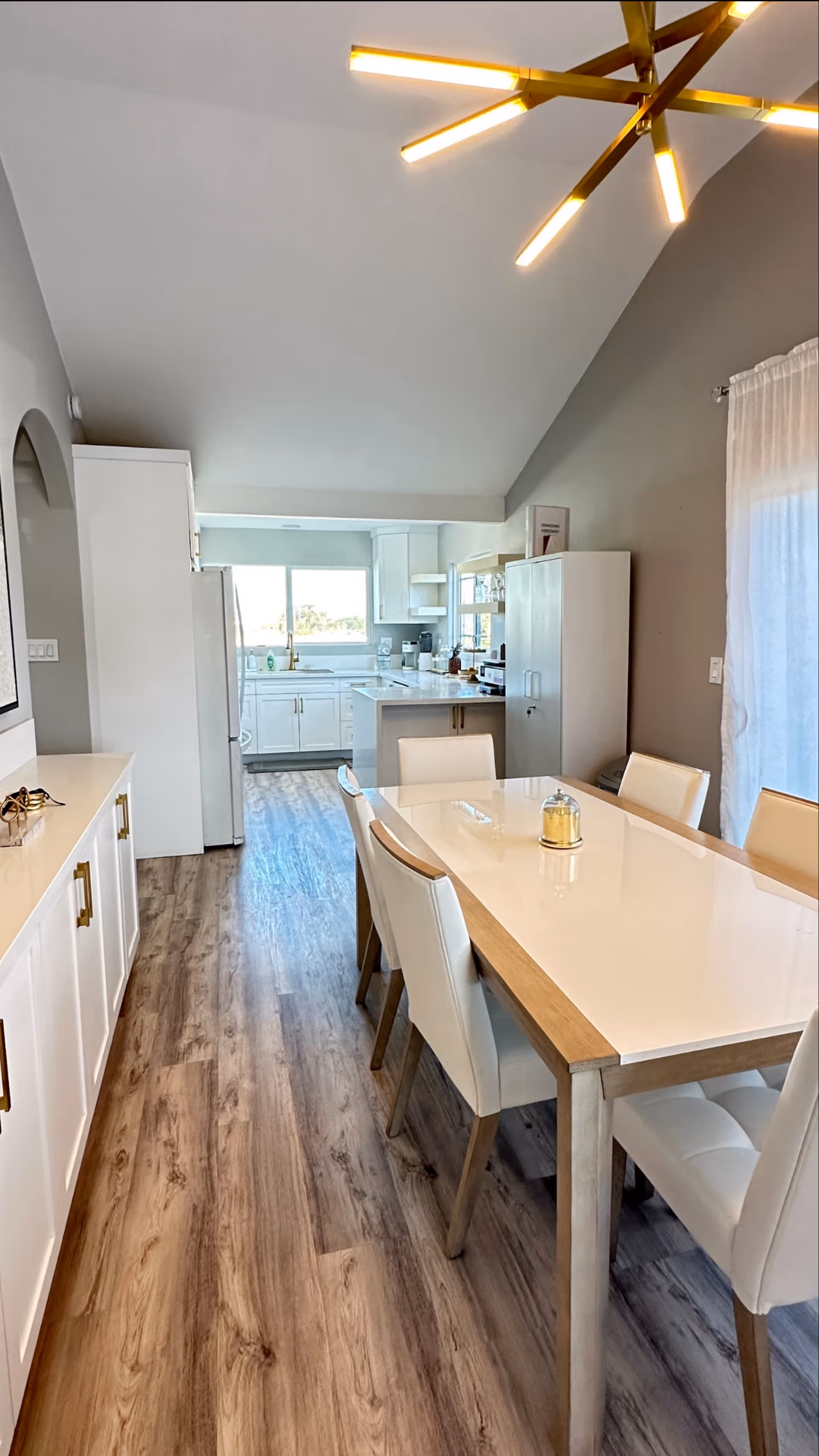 A modern dining area with a white table and six white cushioned chairs. The room has wood flooring and light gray walls. A contemporary gold chandelier hangs from the ceiling. In the background, there is a kitchen area with white cabinets, a refrigerator, and a window letting in natural light.