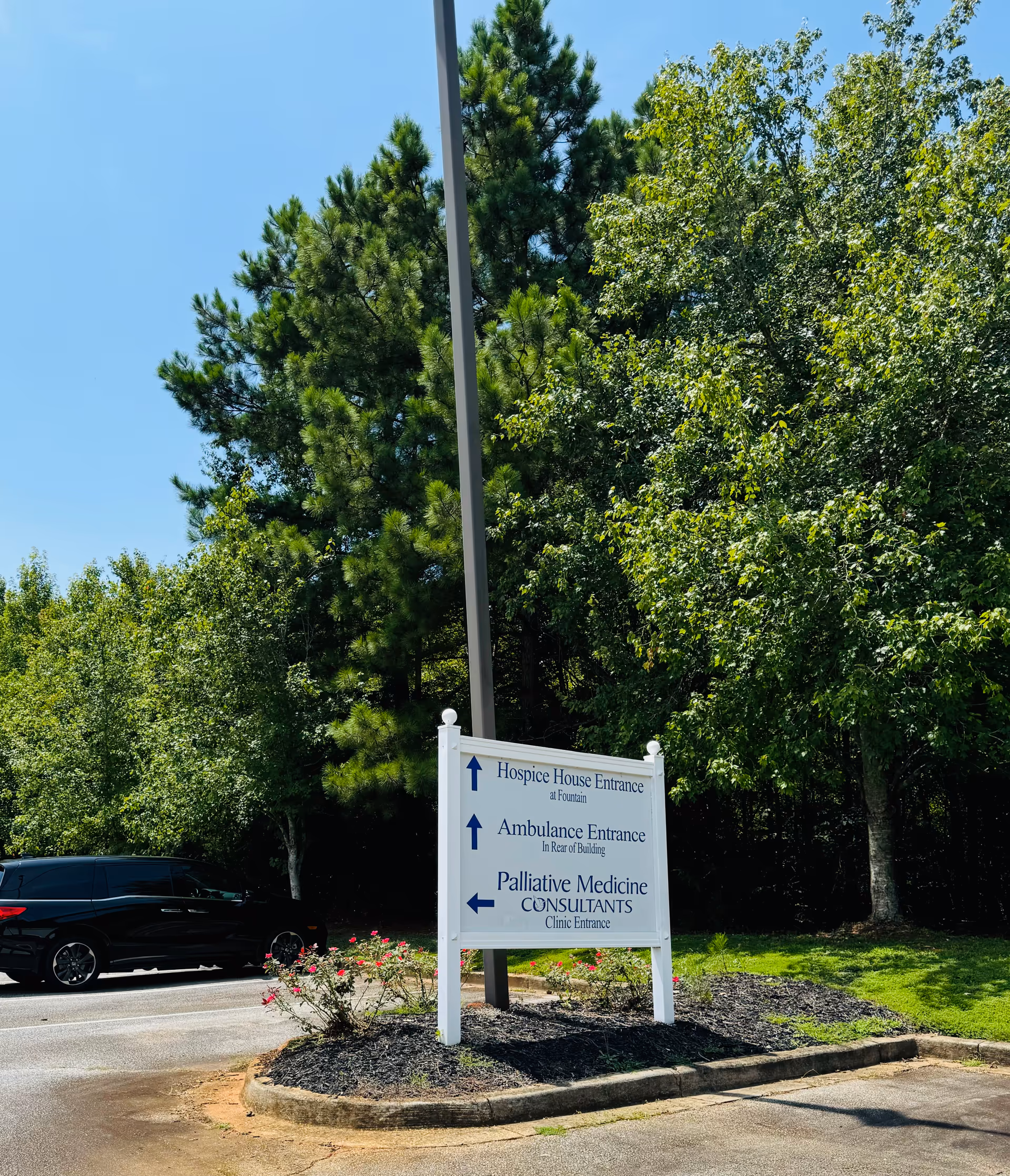 A signboard directs to hospice, ambulance, and palliative care entrances beside parked car.