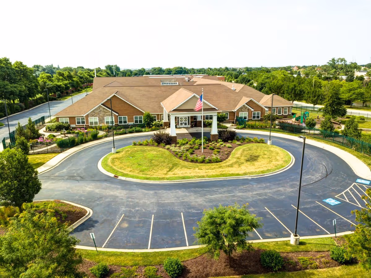 Aerial view of The Auberge at Naperville senior living facility showing a large single-story building with a brown roof and beige exterior. The entrance features a covered porch with white columns and an American flag in front. There is a circular driveway with landscaped greenery in the center and parking spaces surrounding it. Trees and shrubs surround the building and parking area.