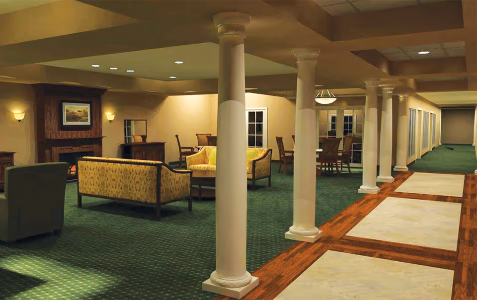 Indoor senior living lounge with green carpet, sofas, chairs, and columns.
