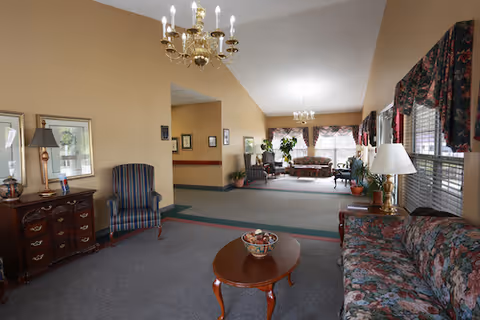 A spacious living room area in a senior living facility with floral patterned sofas, a wooden coffee table with a decorative bowl, a striped armchair, and a wooden chest of drawers with a lamp and framed pictures. The room has beige walls, carpeted floors, large windows with floral curtains, and two chandeliers hanging from the ceiling.