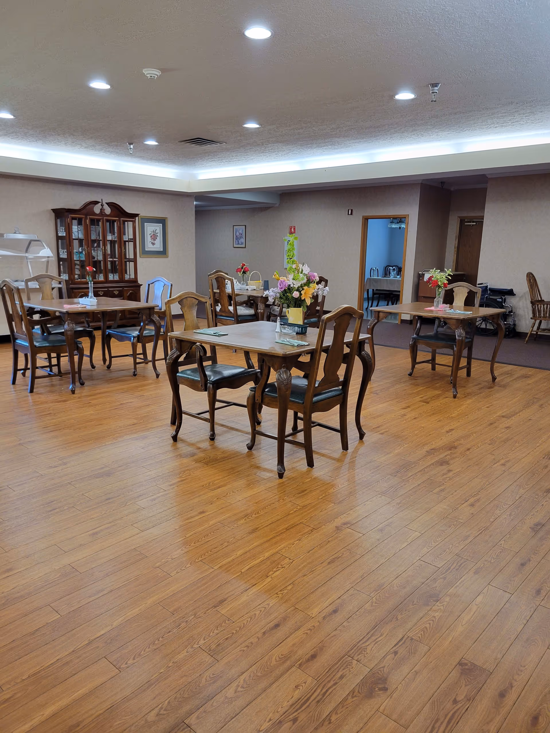 A spacious dining room with wooden floors and several wooden tables and chairs arranged neatly. Each table has a small floral centerpiece. The room has soft lighting from ceiling fixtures and a china cabinet against one wall. There are framed pictures on the walls and an open doorway leading to another room.