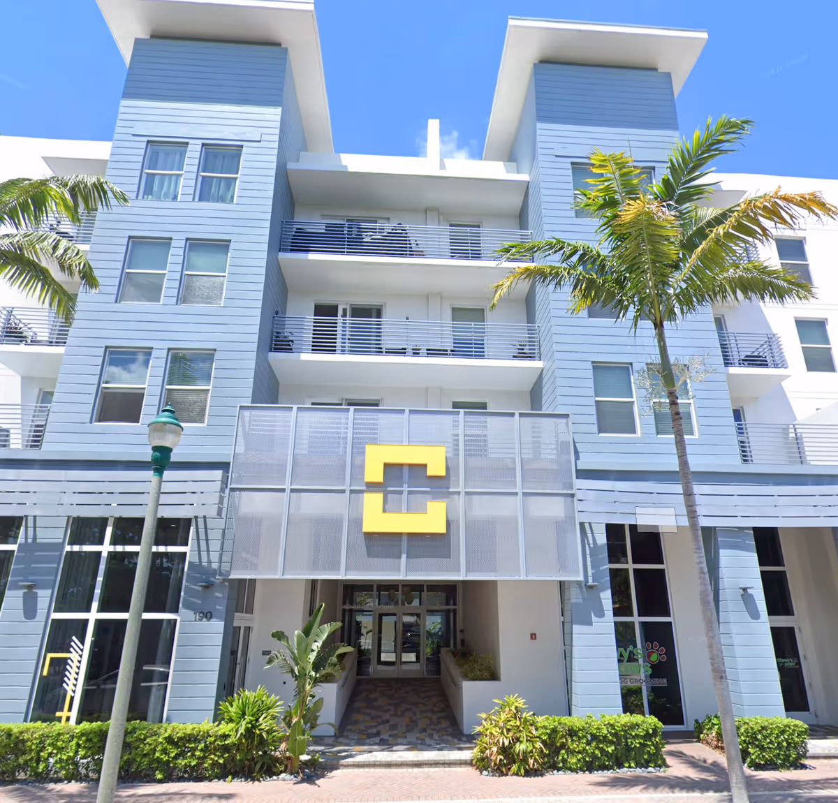 Modern blue apartment building with balconies, palm trees, and a large yellow C sign.